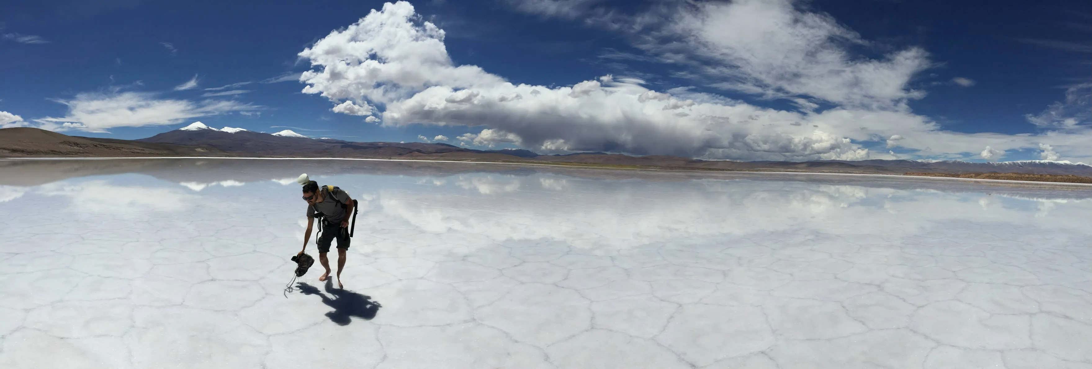 A person bending over survey gear on a flooded salt flat that mirrors the clouds, with snowy peaks on the horizon